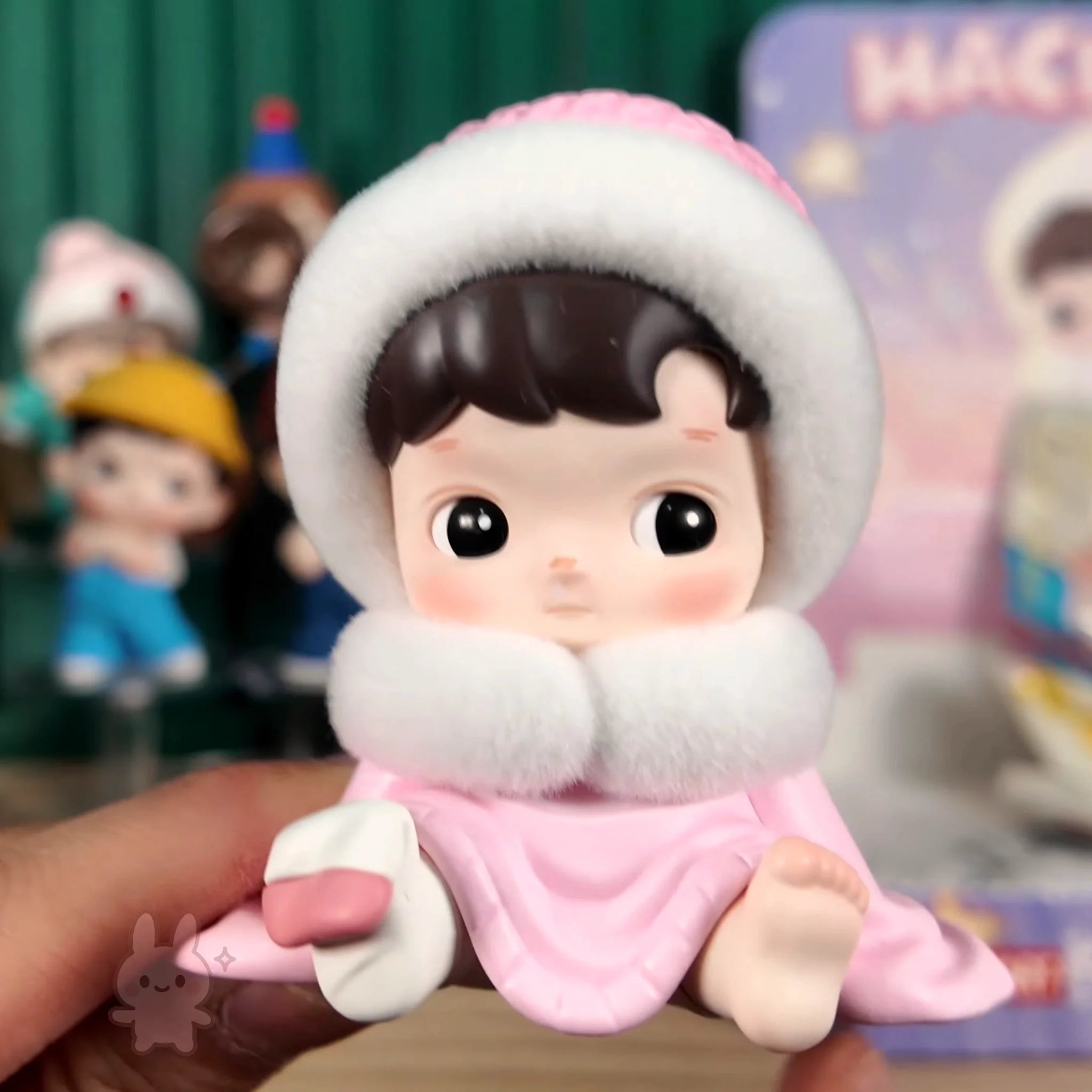 HACIPUPU-Whispers-of-My-World Toy figure of a child in a pink outfit with a white hood held by a hand, with other toys in the background.