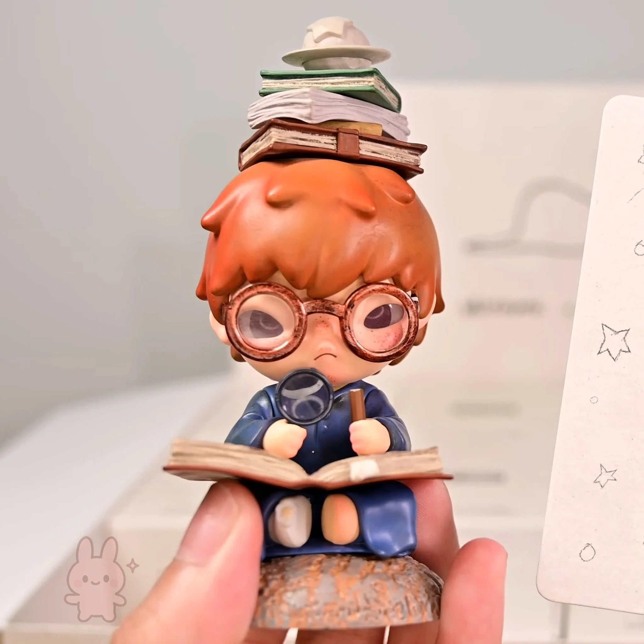 Hirono-Le-Petit-Prince Figurine of a character with glasses and books, held by a hand against a light background.