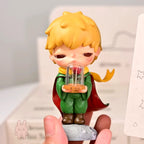 Hirono-Le-Petit-Prince small figurine of a boy with blonde hair and a red cape holding a glass dome with a rose inside, held by a hand against a light background.