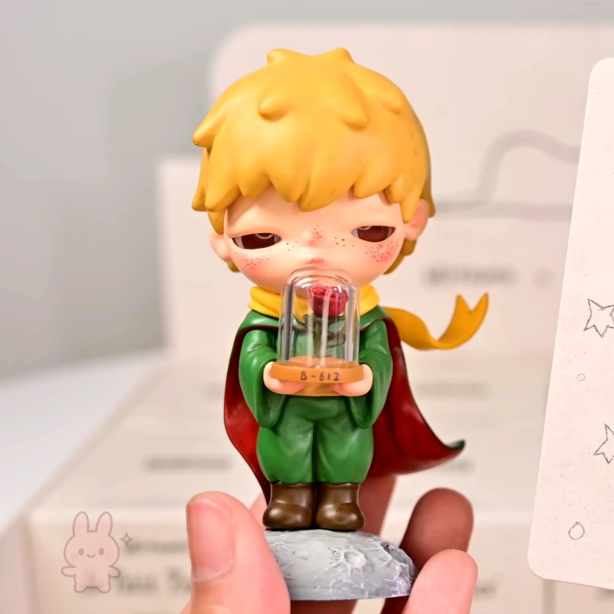 Hirono-Le-Petit-Prince small figurine of a boy with blonde hair and a red cape holding a glass dome with a rose inside, held by a hand against a light background.