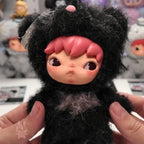 Hirono Road Journal Series Plush Doll Small plush toy with black fur and pink hair held by a person, with a blurred background of other toys.