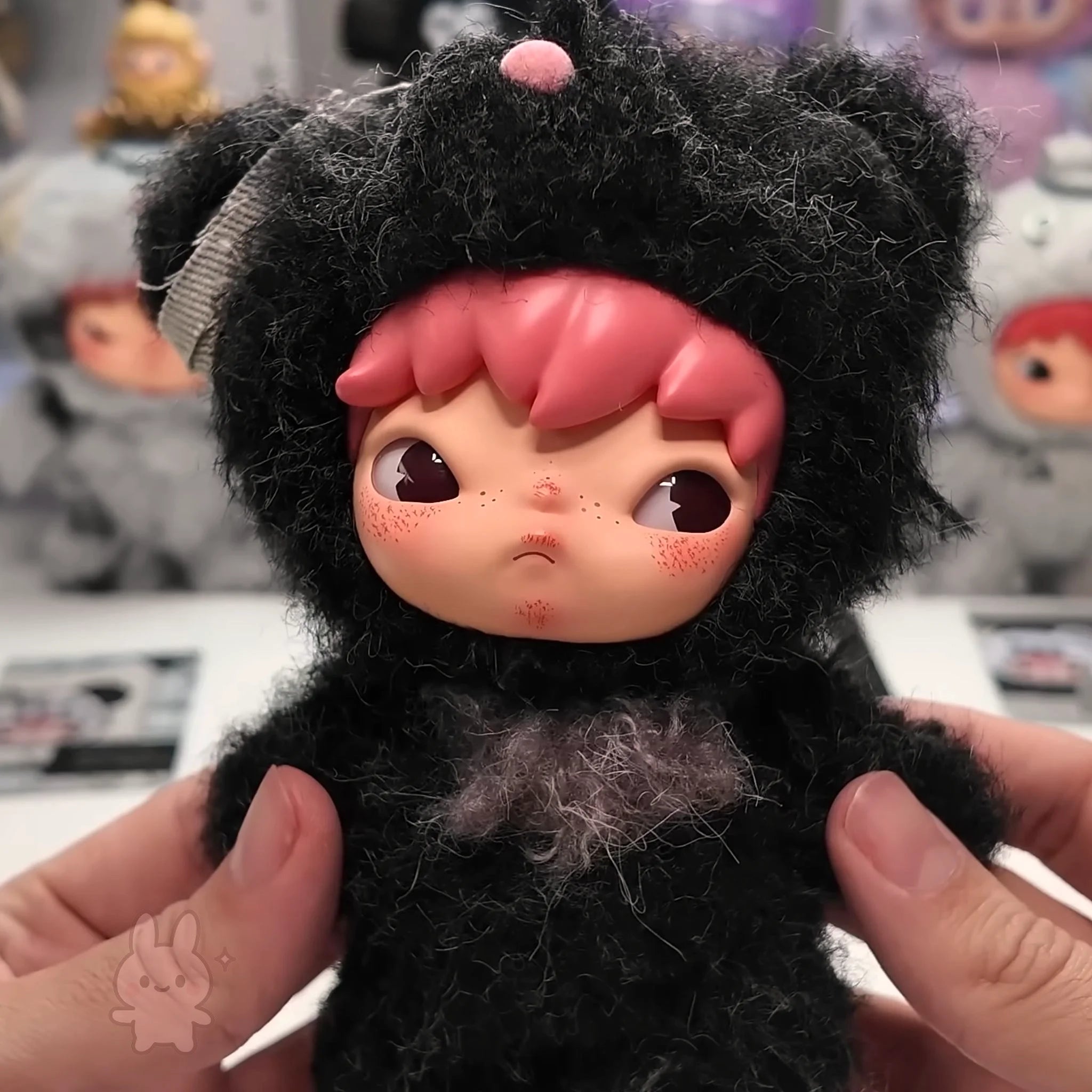 Hirono Road Journal Series Plush Doll Small plush toy with black fur and pink hair held by a person, with a blurred background of other toys.