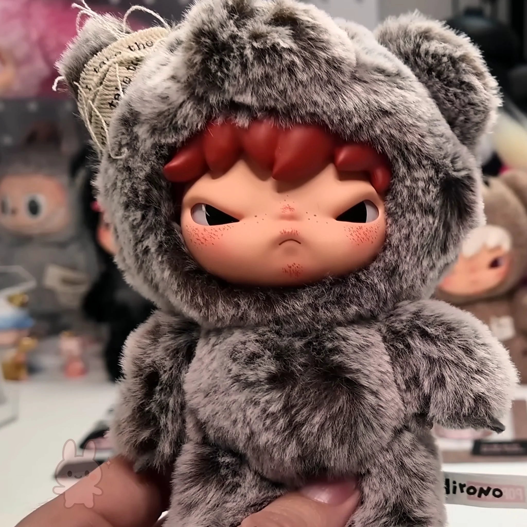 Hirono Road Journal Series Plush Doll Plush toy with a gray furry hood and red face held by a hand, with a blurred background of other toys.