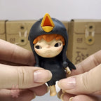 Hirono The Other One Small figurine of a character with a helmet held between two fingers, with a cardboard box in the background.
