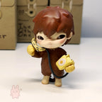 Hirono The Other One Small figurine of a character with brown hair and a yellow object, standing on a white surface with a cardboard box in the background.