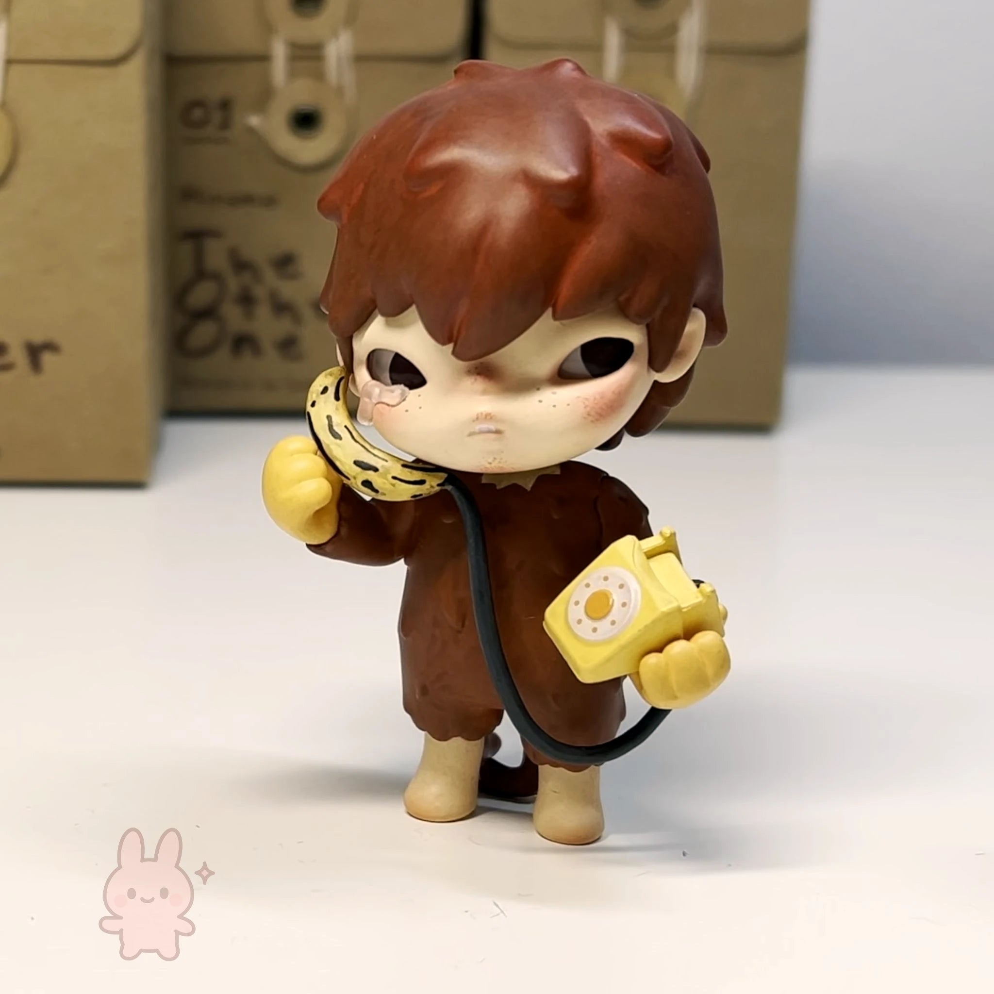 Hirono The Other One Small figurine of a character with brown hair and a yellow object, standing on a white surface with a cardboard box in the background.