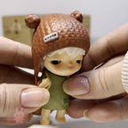 Hirono The Other One Small figurine with brown hat and green outfit held by hands against a neutral background