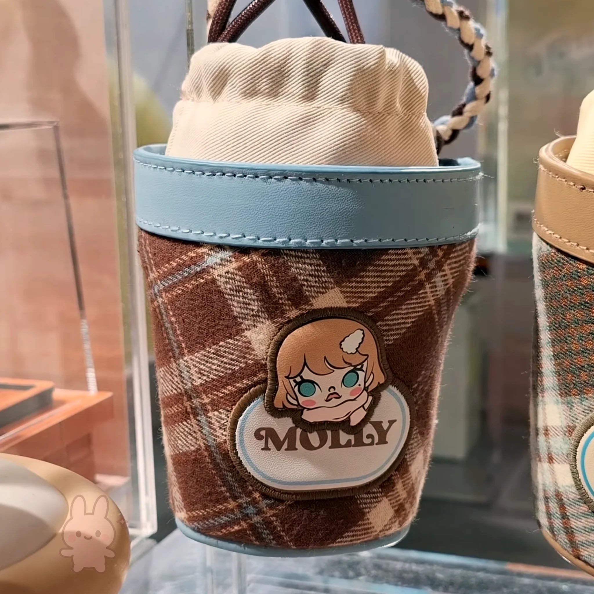 Molly Winter Fit Check Mini Bag Plush toy with a brown checkered sleeve featuring a character named 'Molly' on a glass surface.