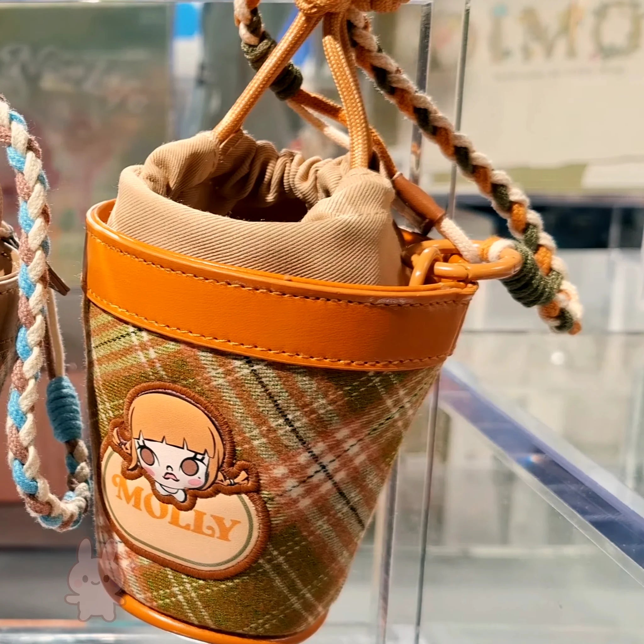 Molly Winter Fit Check Mini Bag Plaid cup holder with orange trim and cartoon character label on a transparent stand.