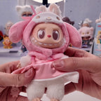 Labubu THE MONSTERS Sanrio Plush toy with pink outfit and bunny ears held by hands against a blurred background of other toys.