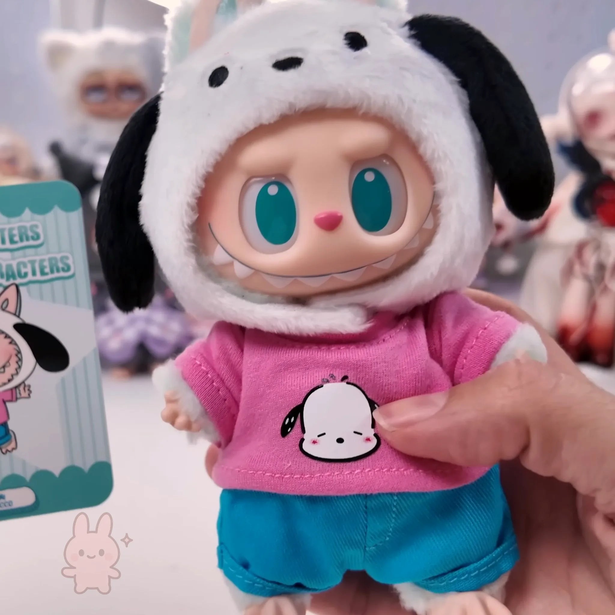 Labubu THE MONSTERS Sanrio Toy figure of a character in a dog costume with a pink shirt and blue shorts, held by a hand.