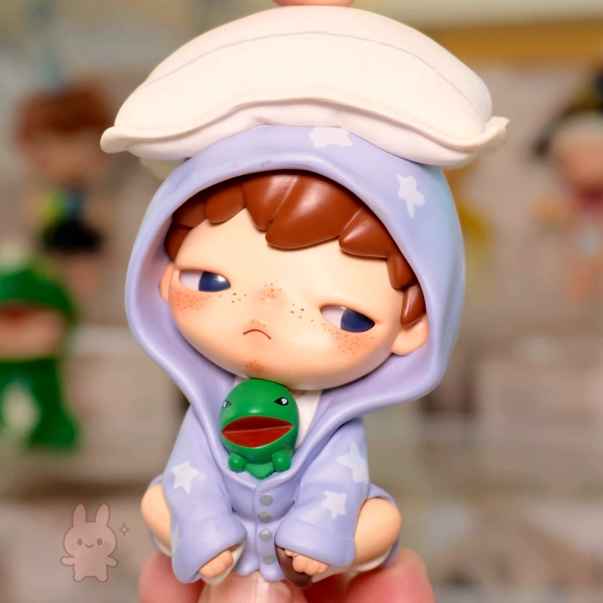Pop Mart Hirono Echo Small figurine of a character wearing a purple hood with a green emblem, held in a hand against a blurred background.
