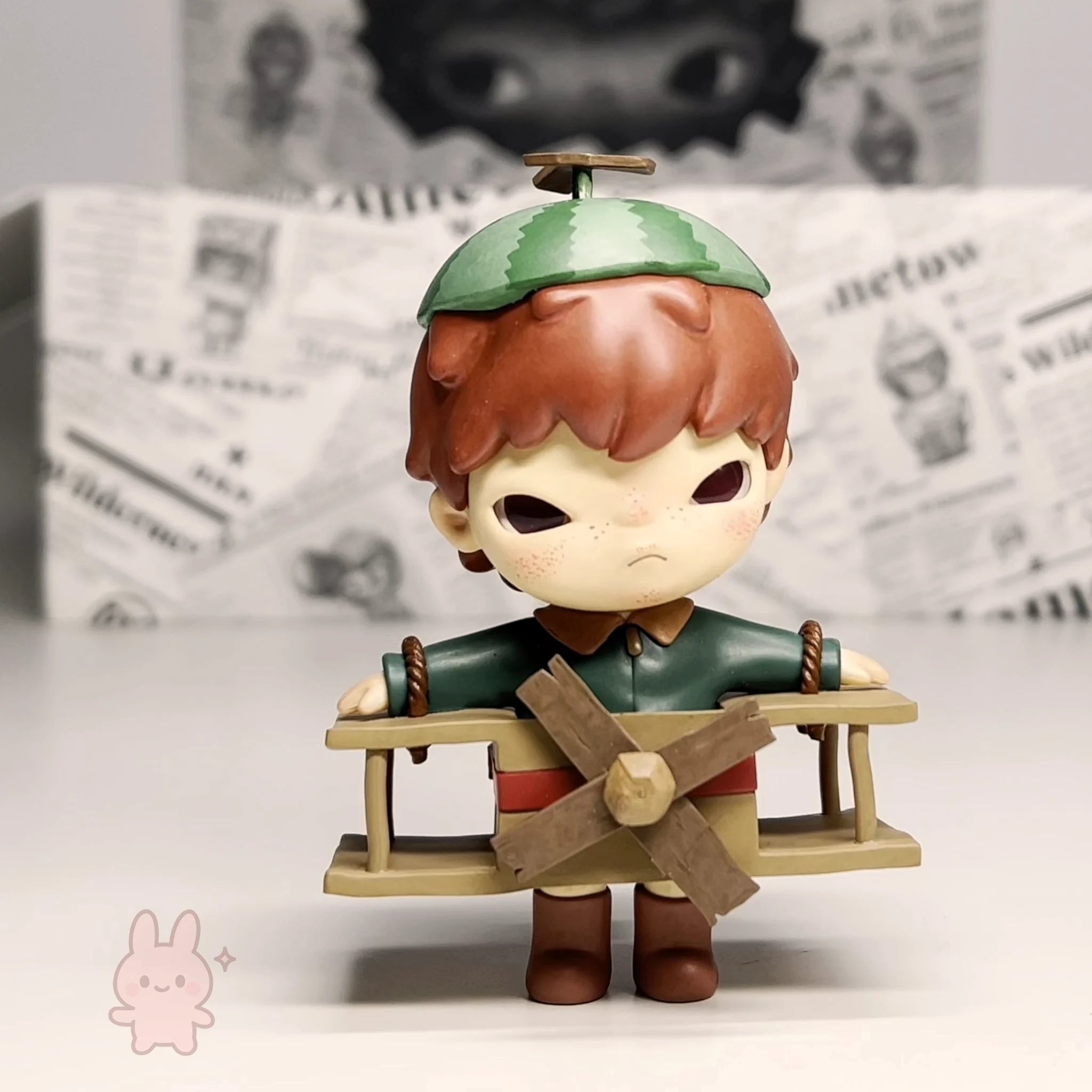 Pop Mart Hirono Little Mischief Small figurine of a character with a green hat and windmill, standing on a white surface with a newspaper background.