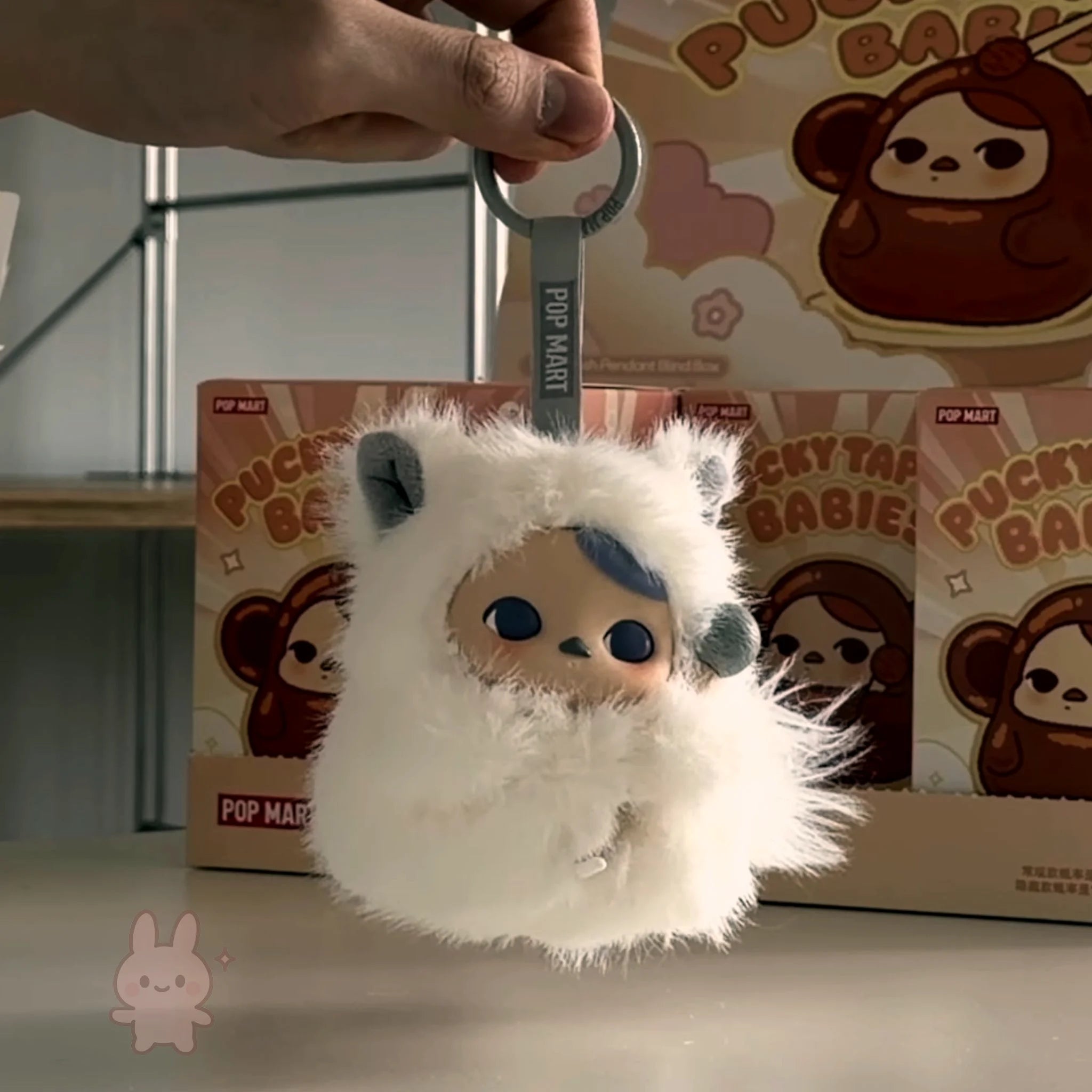 Pop Mart PUCKY Tap Tap Babies White fluffy keychain with blue accents held by a hand, with 'Pucky Baby' packaging in the background.