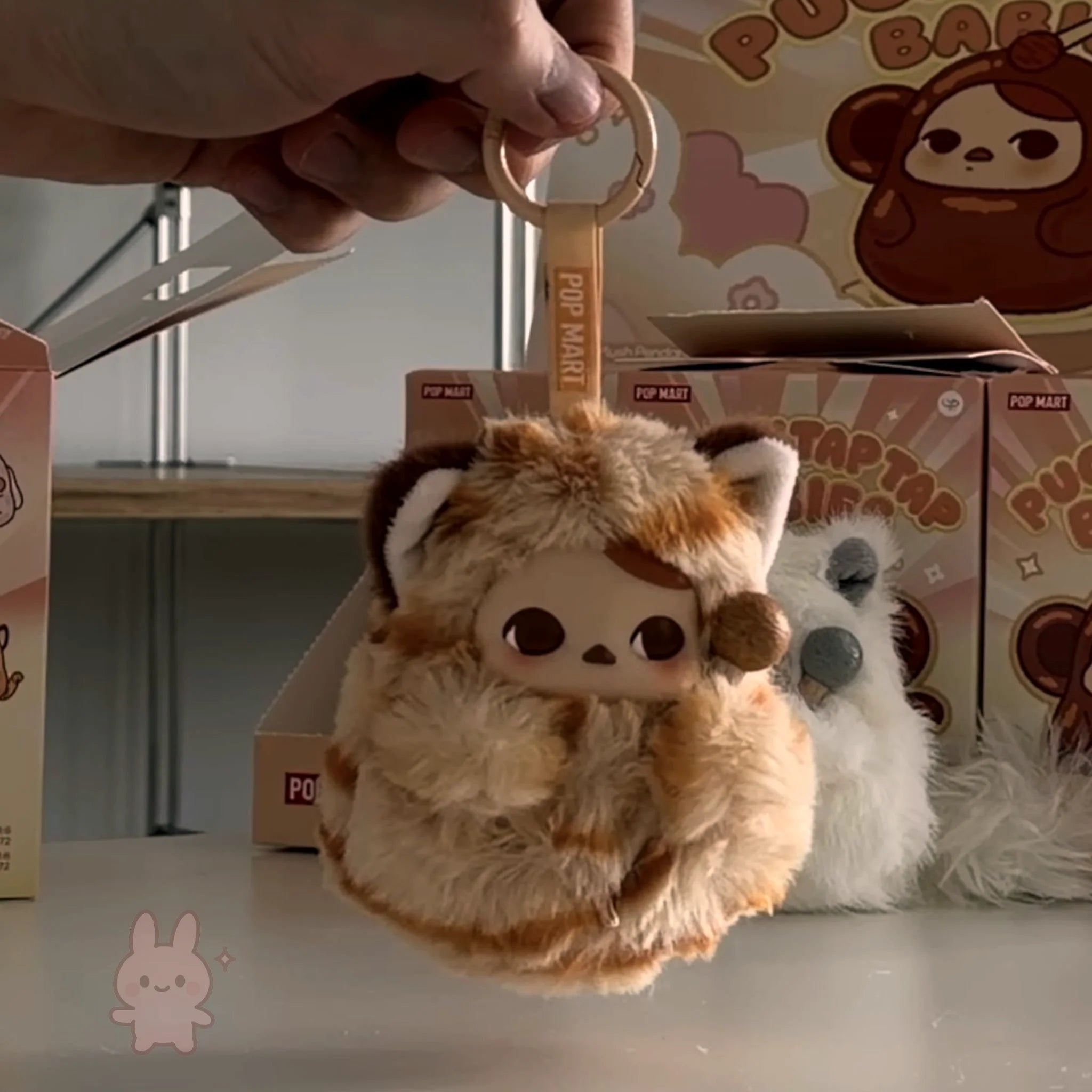 Pop Mart PUCKY Tap Tap Babies Plush keychain with cartoon character held by a hand, with boxes in the background