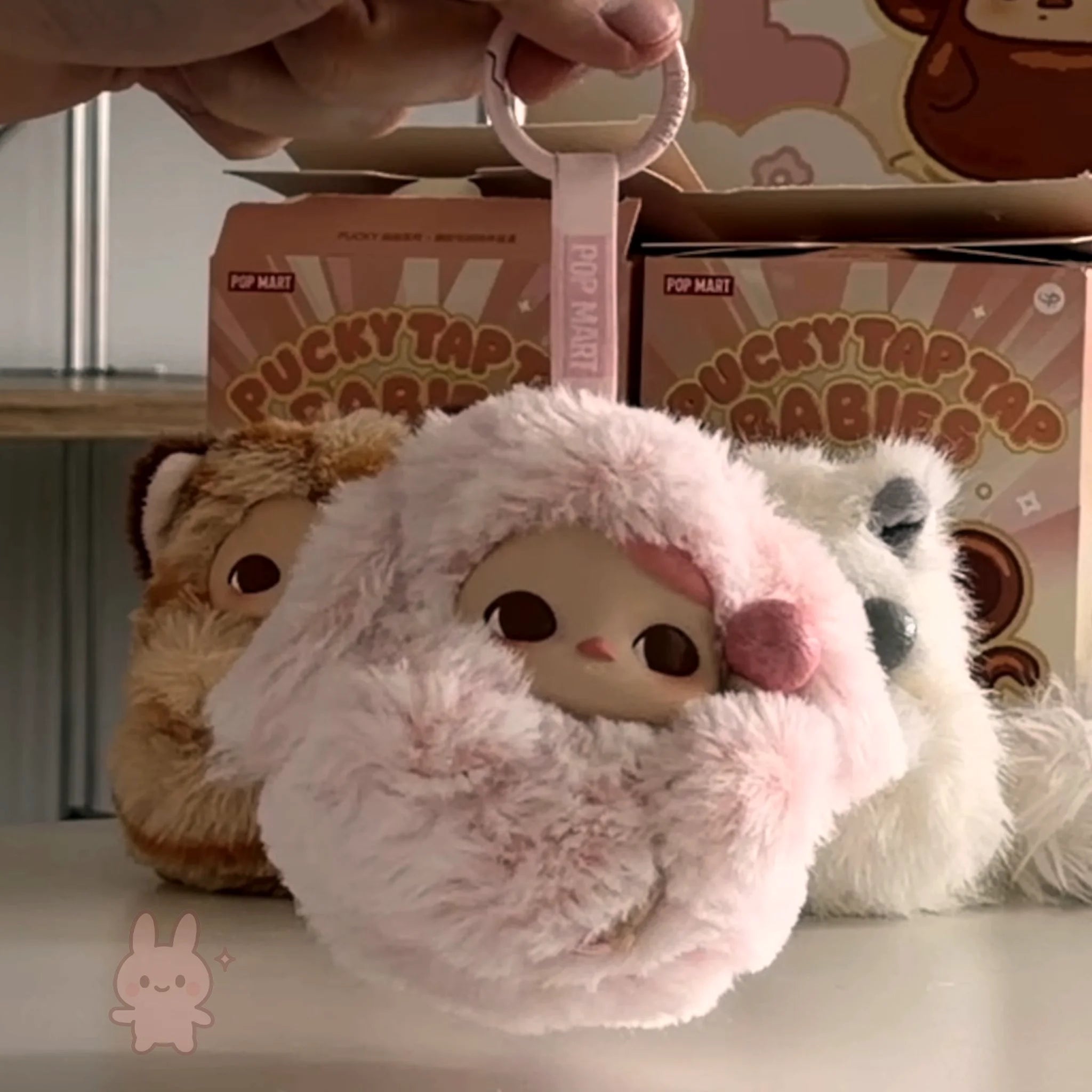 Pop Mart PUCKY Tap Tap Babies Plush toy with pink fluffy hood held by a hand, with 'Pucky Tap Tap' boxes in the background.