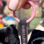 Skullpanda x My Melody Pink keychain with 'OSKULLPANIC' branding held by a hand against a blurred background.