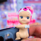 Sunny Angel Hippers Toy figure of a baby with a pink helmet held by a hand against a colorful blurred background