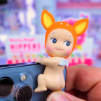 Sunny Angel Hippers Toy figure with orange head and blue eyes held by a hand against a colorful blurred background