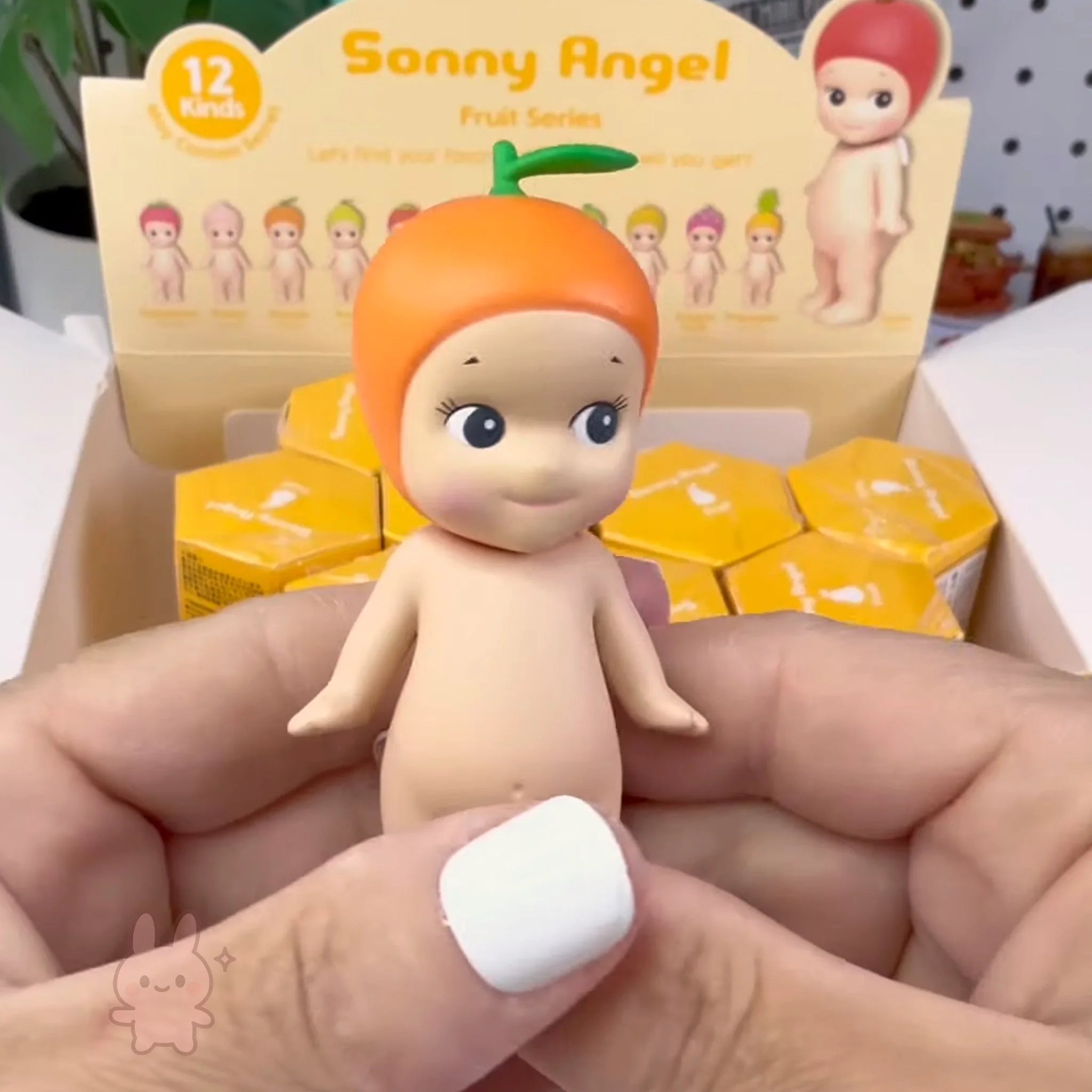 Sonny Angel fruit Toy figure with orange hair held by a hand, with packaging in the background