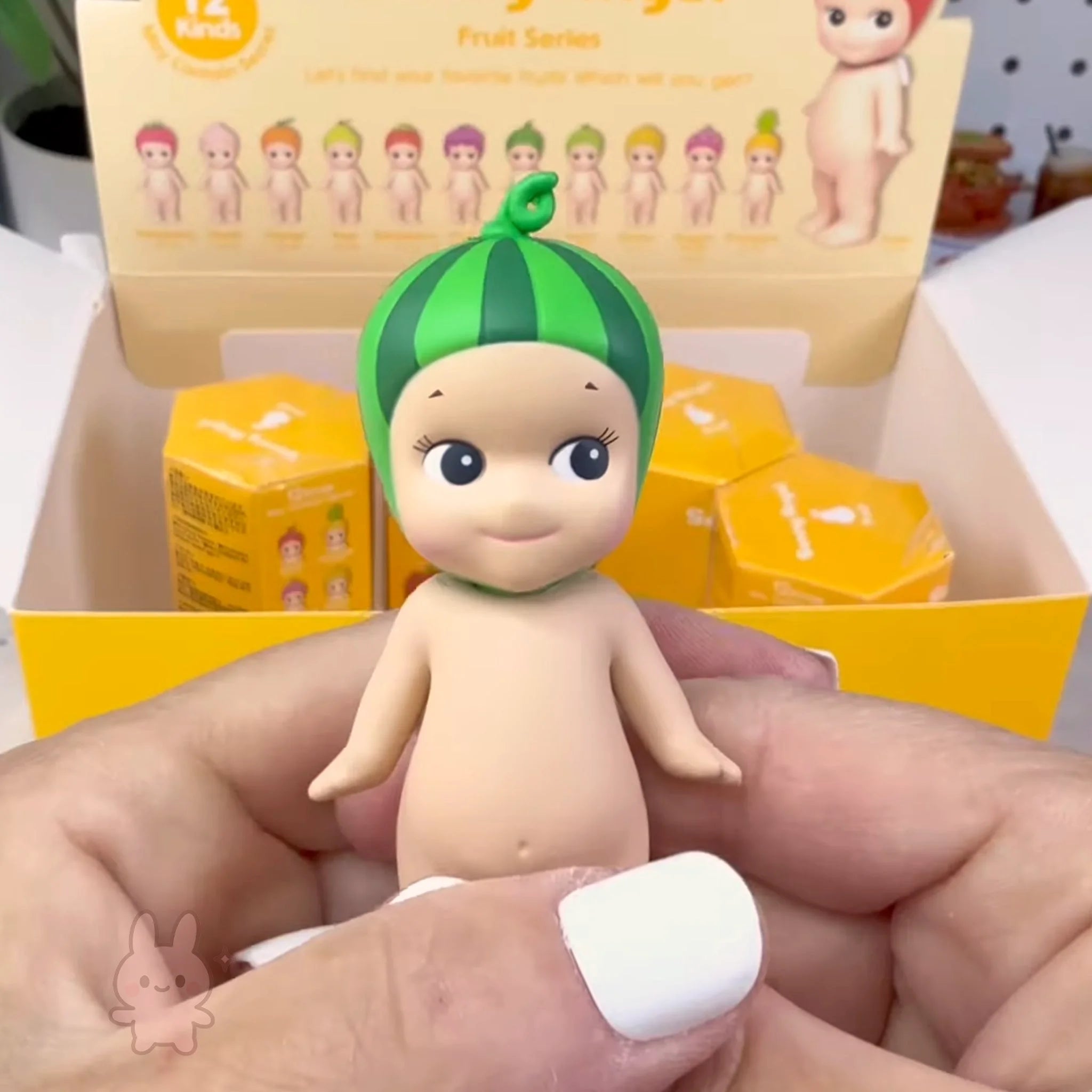 Sonny Angel fruit Toy figure with a watermelon-themed head held by a hand, with packaging in the background.