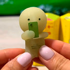 SMISKI Small round character with a green card held by a hand against a blurred background