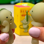 SMISKI Two small green figurines held by hands with pink nail polish, against a blurred background.