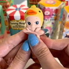 Sonny-Angel-hippers-animal Toy figure of a baby with a duck head held by hands with blue nail polish, colorful boxes in the background.