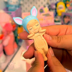 Sonny Angel hippers dreaming Toy figure of a baby with bunny ears held by a hand against a colorful store background