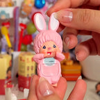 Monchhic hiippers Pink toy figure with bunny ears held by a hand against a colorful background