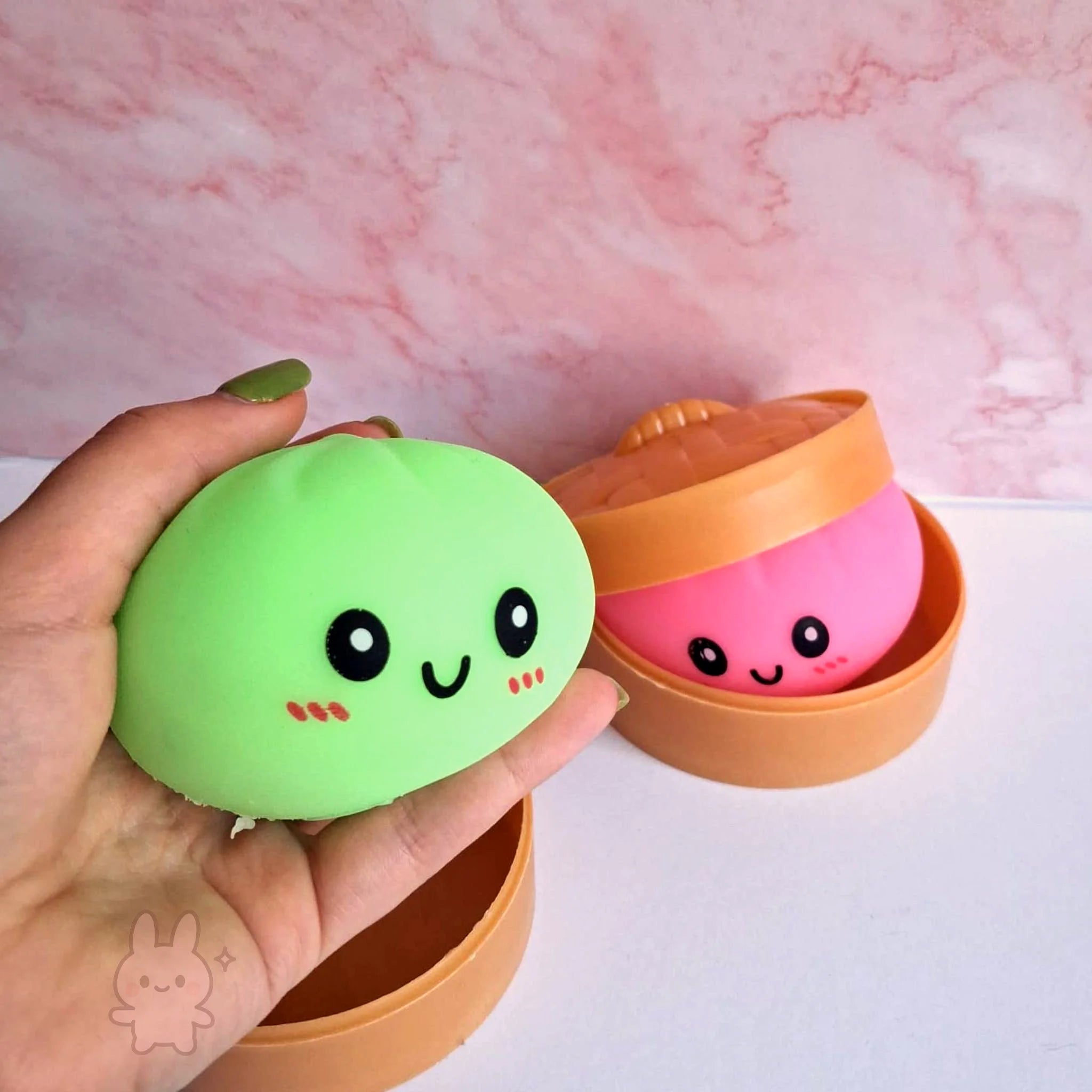 Two cute, round squishies in orange containers held by a hand against a pink marble background.