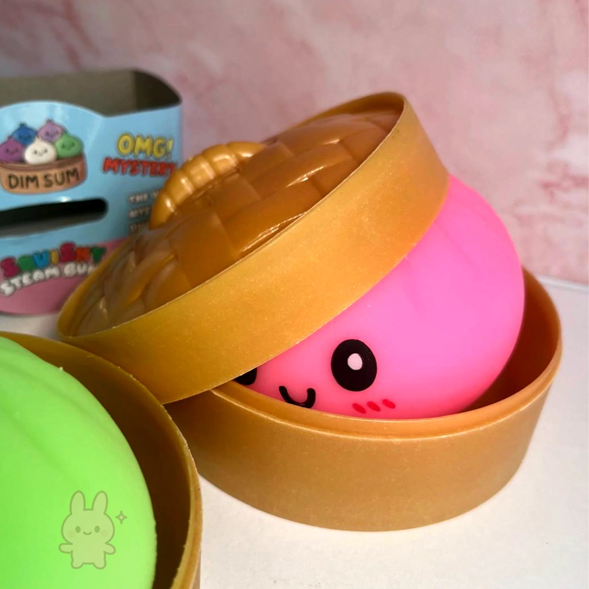 Pink squishy toy with a face inside an open brown box on a pink background.