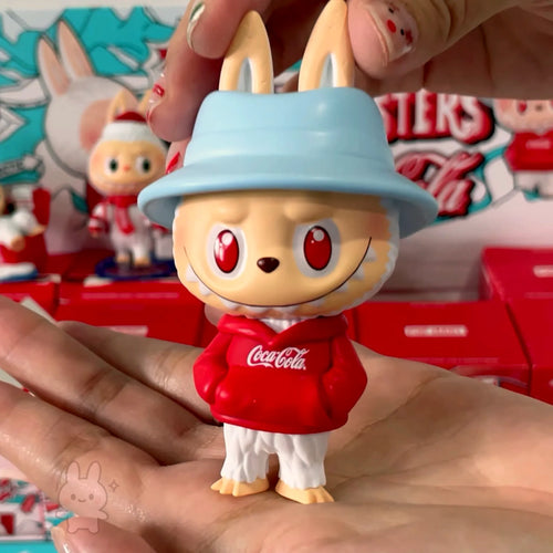 The Monsters Coca-Cola Toy figure of a character wearing a Coca-Cola hoodie and light blue hat, held in a hand with a colorful background.