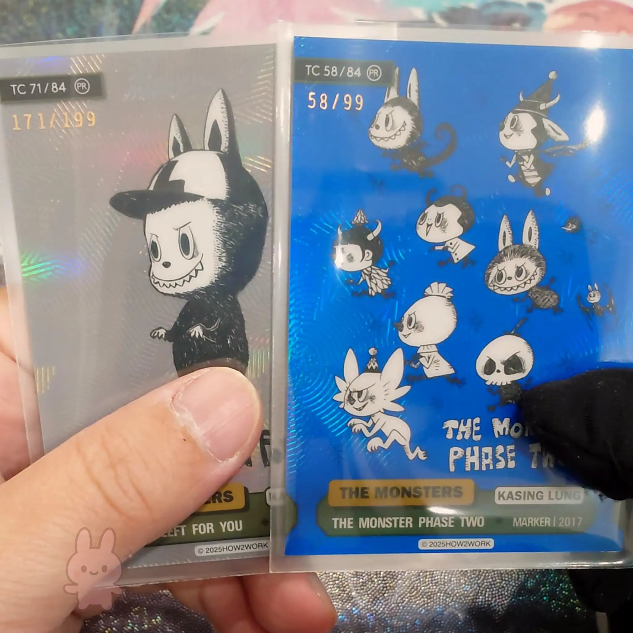 Two trading cards held by a hand, one with a black and white character and the other with multiple small characters on a blue background.