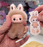Plush toy resembling a character in a bunny costume held by a hand, with a matching sticker.