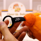Orange plush toy with a 'cry baby' tag held by a hand, with a blurred background.