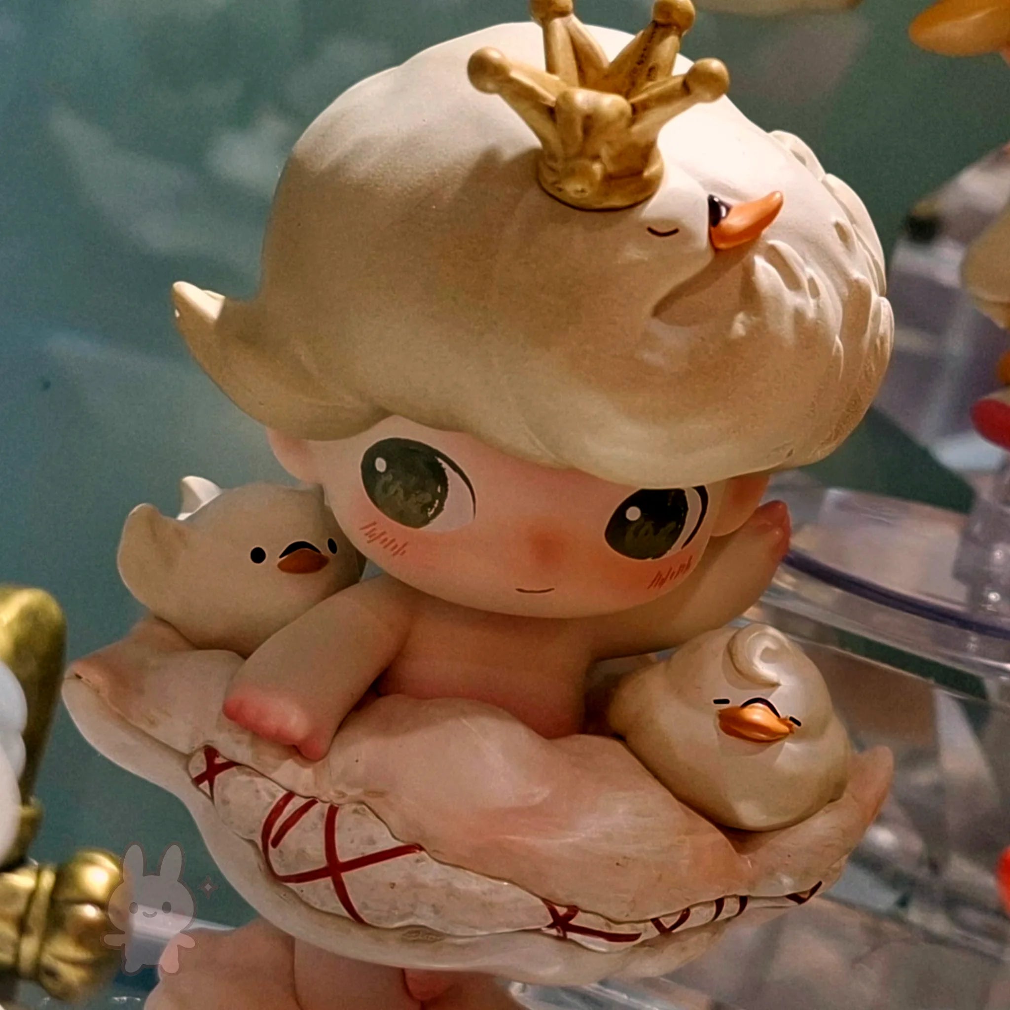 dimoo-the-missing-day Cute figurine of a baby with a crown holding two ducks, on a blue background.