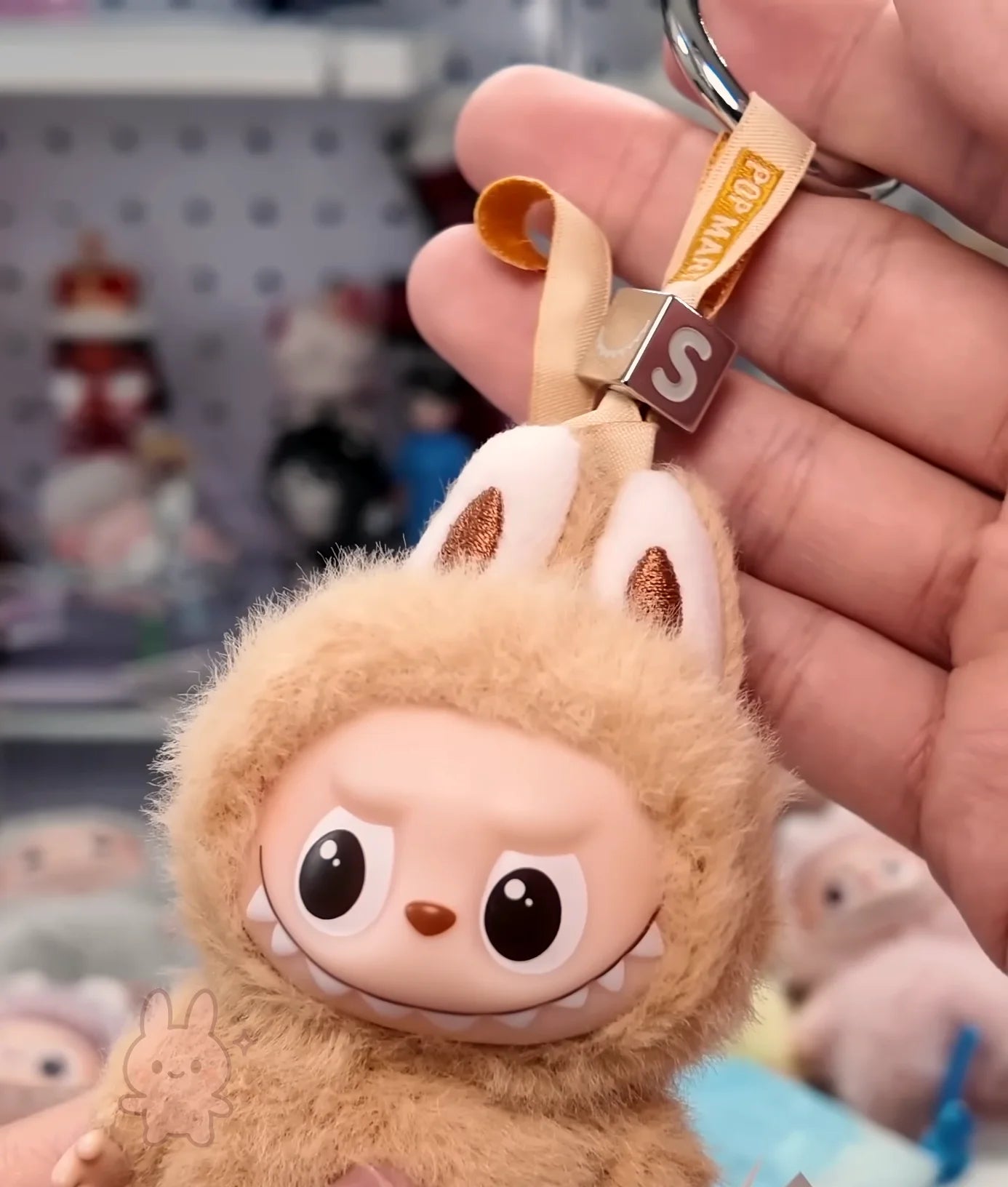 Labubu Plush keychain with a cartoonish design held by a hand against a blurred background of toys.