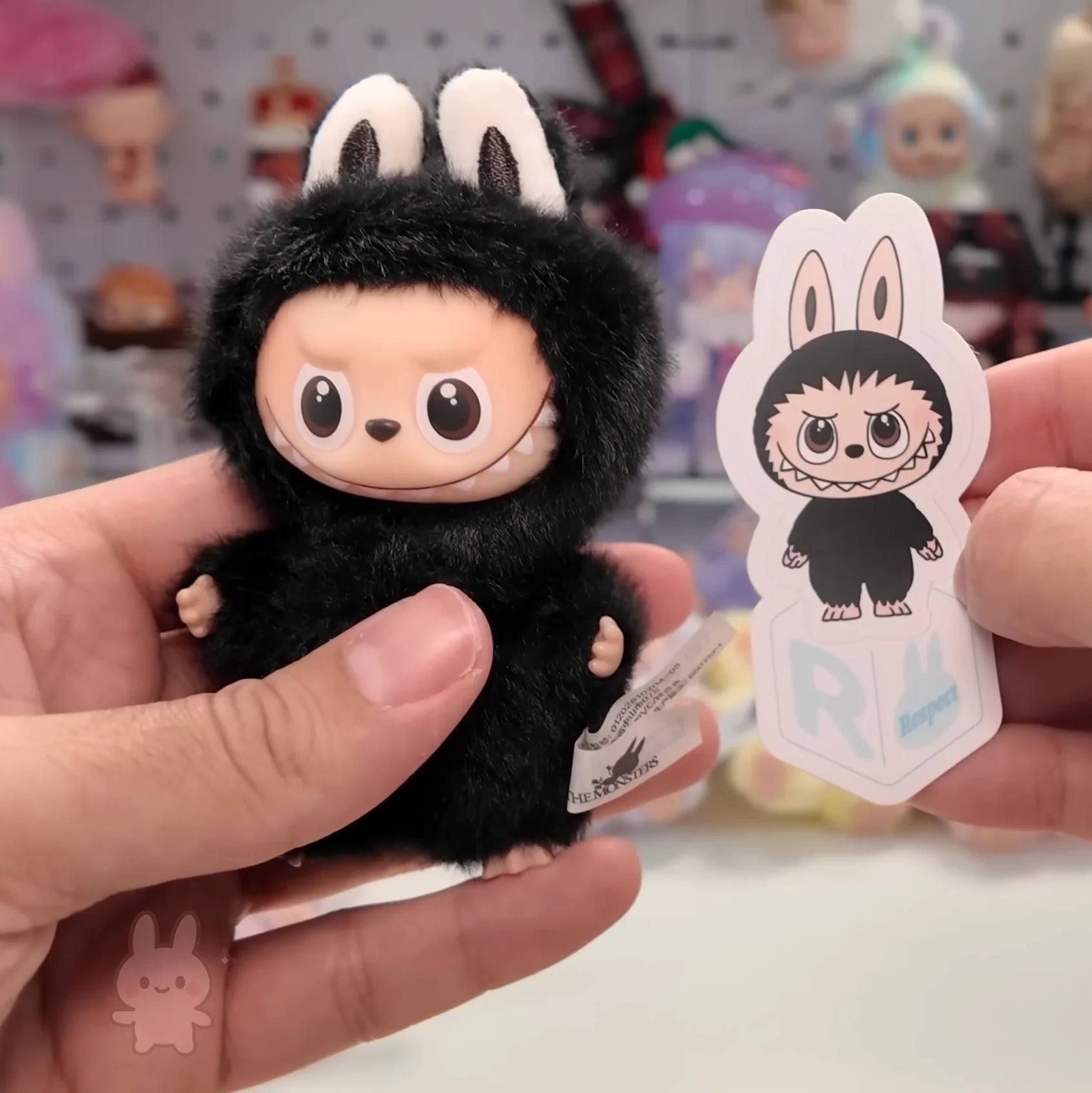 Plush toy with rabbit ears held by a hand, with a sticker of the same character in the background.
