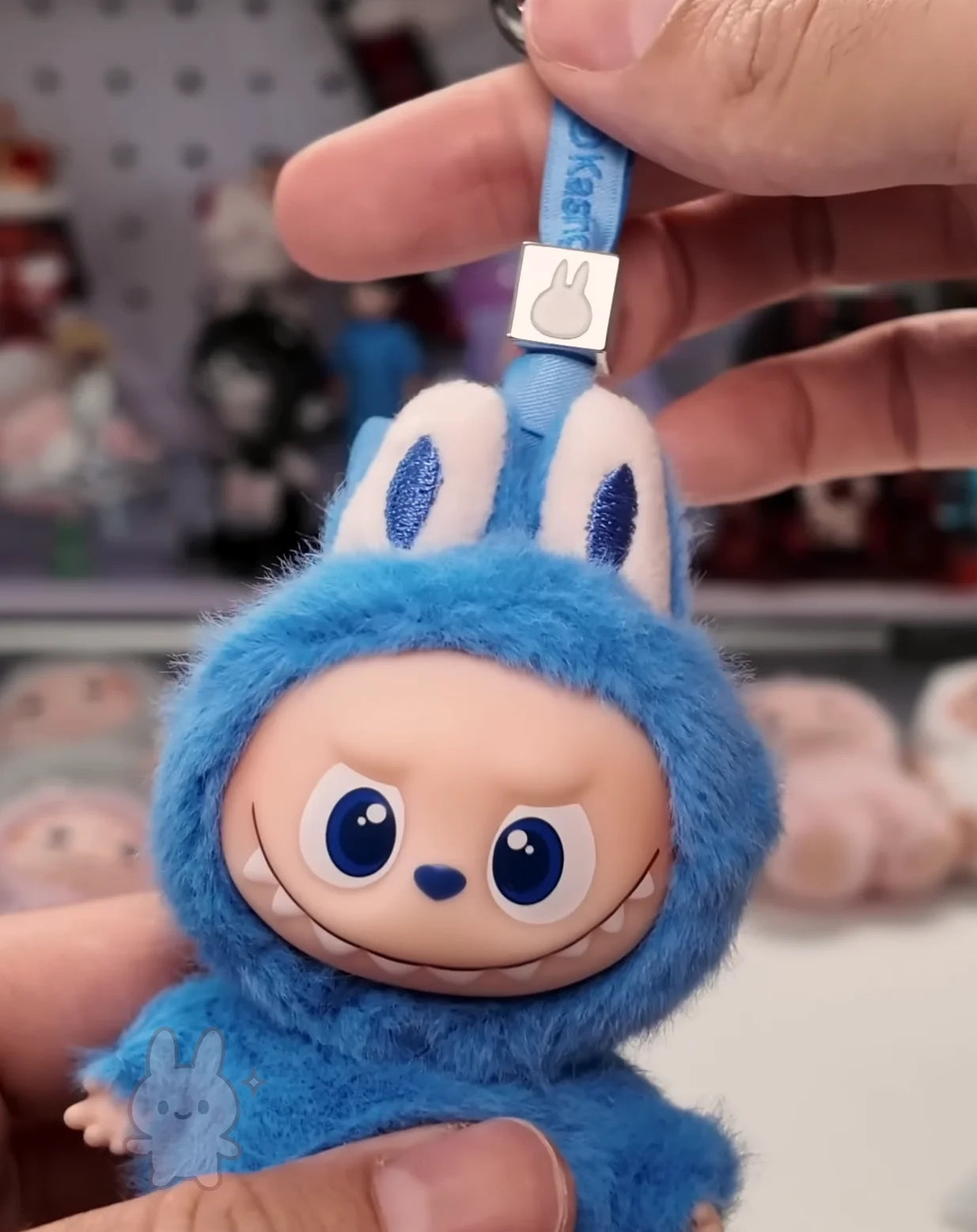 Labubu Blue plush keychain with a smiling face held by a hand against a blurred background
