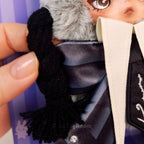 skullpanda Close-up of a hand holding a black knitted scarf over a doll with a striped shirt and coat.