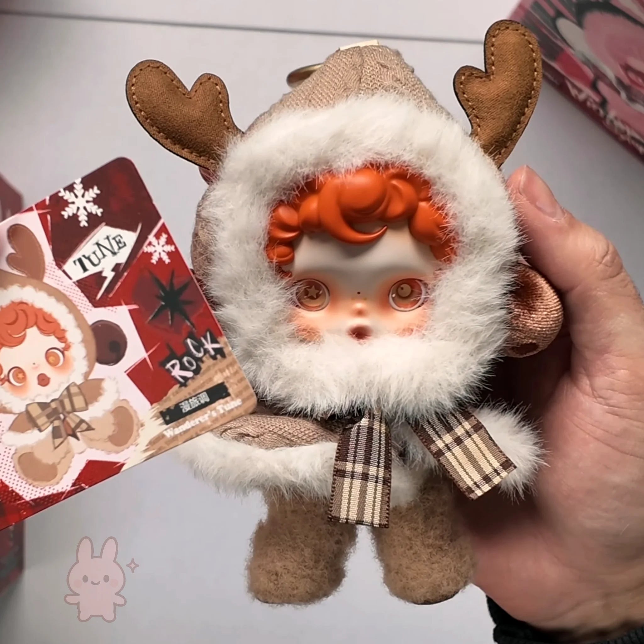 skullpanda Stuffed toy with reindeer antlers and a plaid bow held by a hand, with a blurred background.