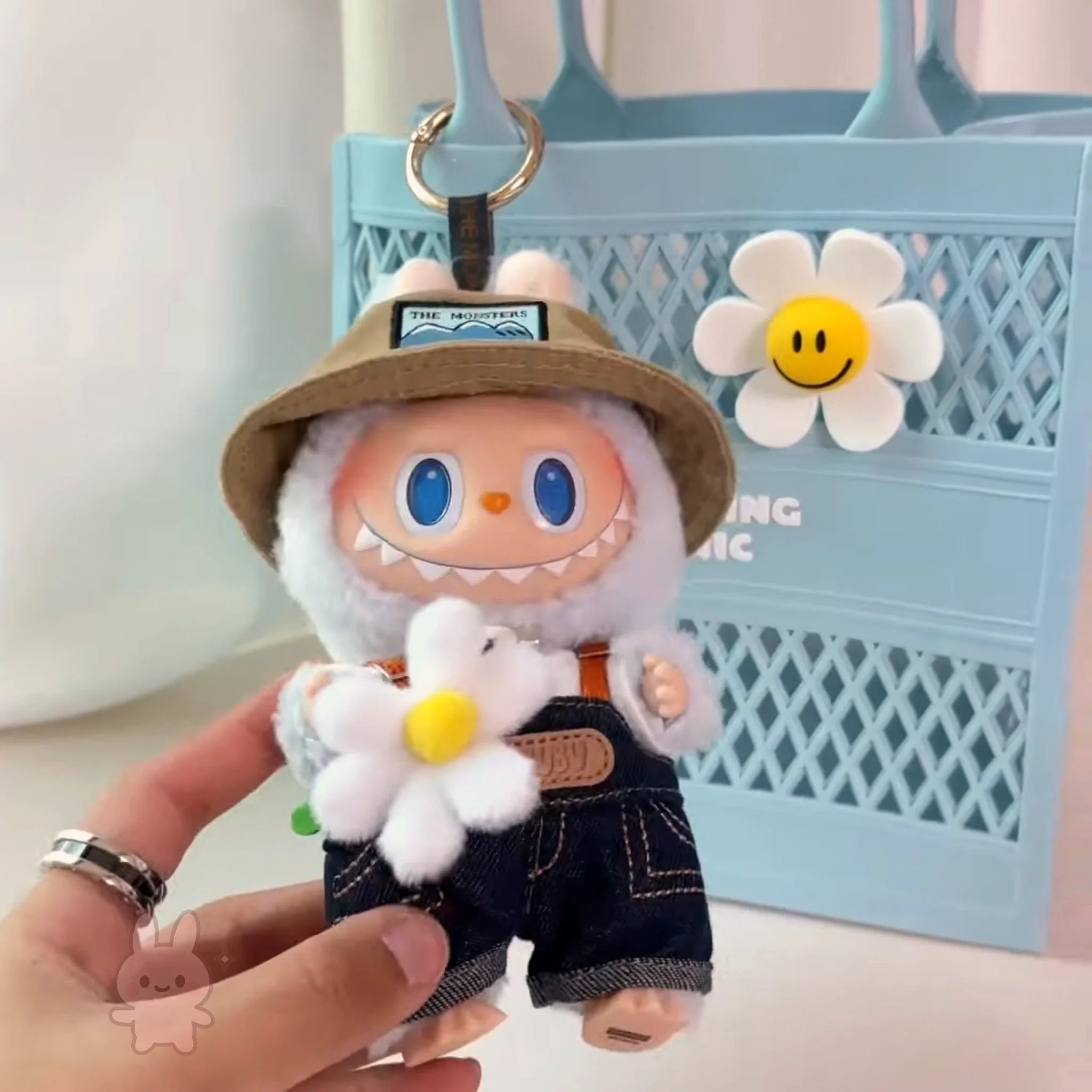 Labubu Plush keychain of a character with a hat and overalls, holding a flower, against a light blue background.
